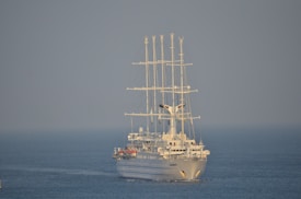 A large white sailing ship with multiple masts is navigating through calm blue waters. The ship appears modern with sleek lines and is equipped with large sails. The sky is clear, contributing to a serene maritime setting.