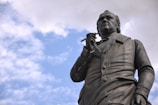 The image features a bronze statue of a historical figure holding a writing instrument. The statue presents the person in an upright and dignified posture, looking slightly upwards. The background is a partly cloudy sky, giving a serene atmosphere.