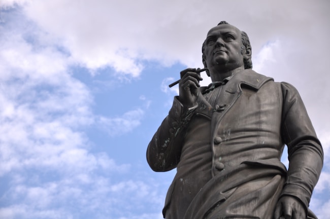 The image features a bronze statue of a historical figure holding a writing instrument. The statue presents the person in an upright and dignified posture, looking slightly upwards. The background is a partly cloudy sky, giving a serene atmosphere.