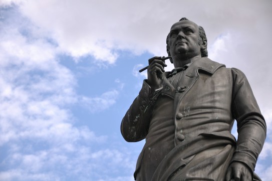 The image features a bronze statue of a historical figure holding a writing instrument. The statue presents the person in an upright and dignified posture, looking slightly upwards. The background is a partly cloudy sky, giving a serene atmosphere.