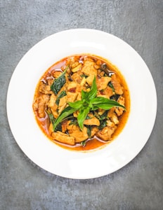 A plate of stir-fried dish consisting of pieces of meat, possibly chicken, garnished with fresh basil leaves. The food is immersed in a reddish-brown sauce, likely indicative of a spicy or savory flavor. The dish is presented on a white round plate against a textured gray surface.