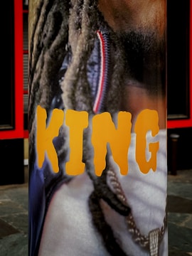 A vertical banner featuring a close-up image of a person with long dreadlocks and a gold chain around their neck. The word 'KING' is prominently displayed in bold, yellow letters across the image.