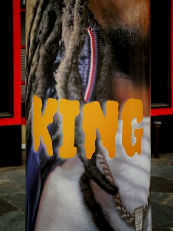A vertical banner featuring a close-up image of a person with long dreadlocks and a gold chain around their neck. The word 'KING' is prominently displayed in bold, yellow letters across the image.