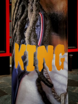 A vertical banner featuring a close-up image of a person with long dreadlocks and a gold chain around their neck. The word 'KING' is prominently displayed in bold, yellow letters across the image.