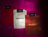 Three folded ribbed shirts are neatly arranged on a bright magenta furry surface. Each shirt is labeled with a 'MANGO' brand tag. The shirts are in red, white, and brown colors, with the word 'MANGO' printed on them in contrasting thread colors.