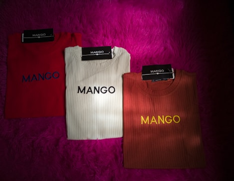 Three folded ribbed shirts are neatly arranged on a bright magenta furry surface. Each shirt is labeled with a 'MANGO' brand tag. The shirts are in red, white, and brown colors, with the word 'MANGO' printed on them in contrasting thread colors.