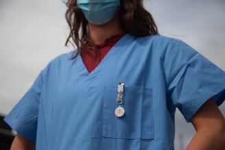 A person wearing a blue medical scrub top and a face mask. The scrub has a badge or ID clipped to the pocket. The background shows a clear sky.