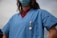 A person wearing a blue medical scrub top and a face mask. The scrub has a badge or ID clipped to the pocket. The background shows a clear sky.