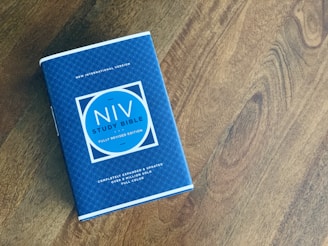 A blue NIV Study Bible is placed on a wooden surface. The book cover features a geometric pattern with a bold circular design containing the title and edition information. The Bible is slightly tilted and positioned towards the left side of the image.