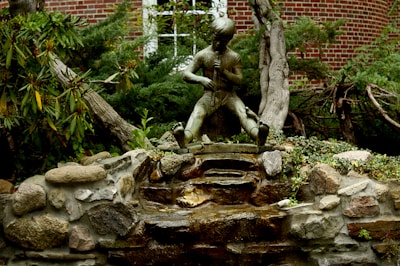 A bronze statue of a young boy kneeling and playing a flute is situated in a garden setting. The statue is mounted on a rock formation that resembles a small waterfall. Surrounding the statue are lush green bushes and trees, with a brick building visible in the background.