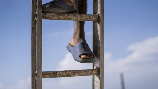 A close-up view of a person's foot wearing a dark sandal while stepping onto a metal ladder against a blue sky backdrop. The person's shin and toes are visible, displaying a relaxed posture.
