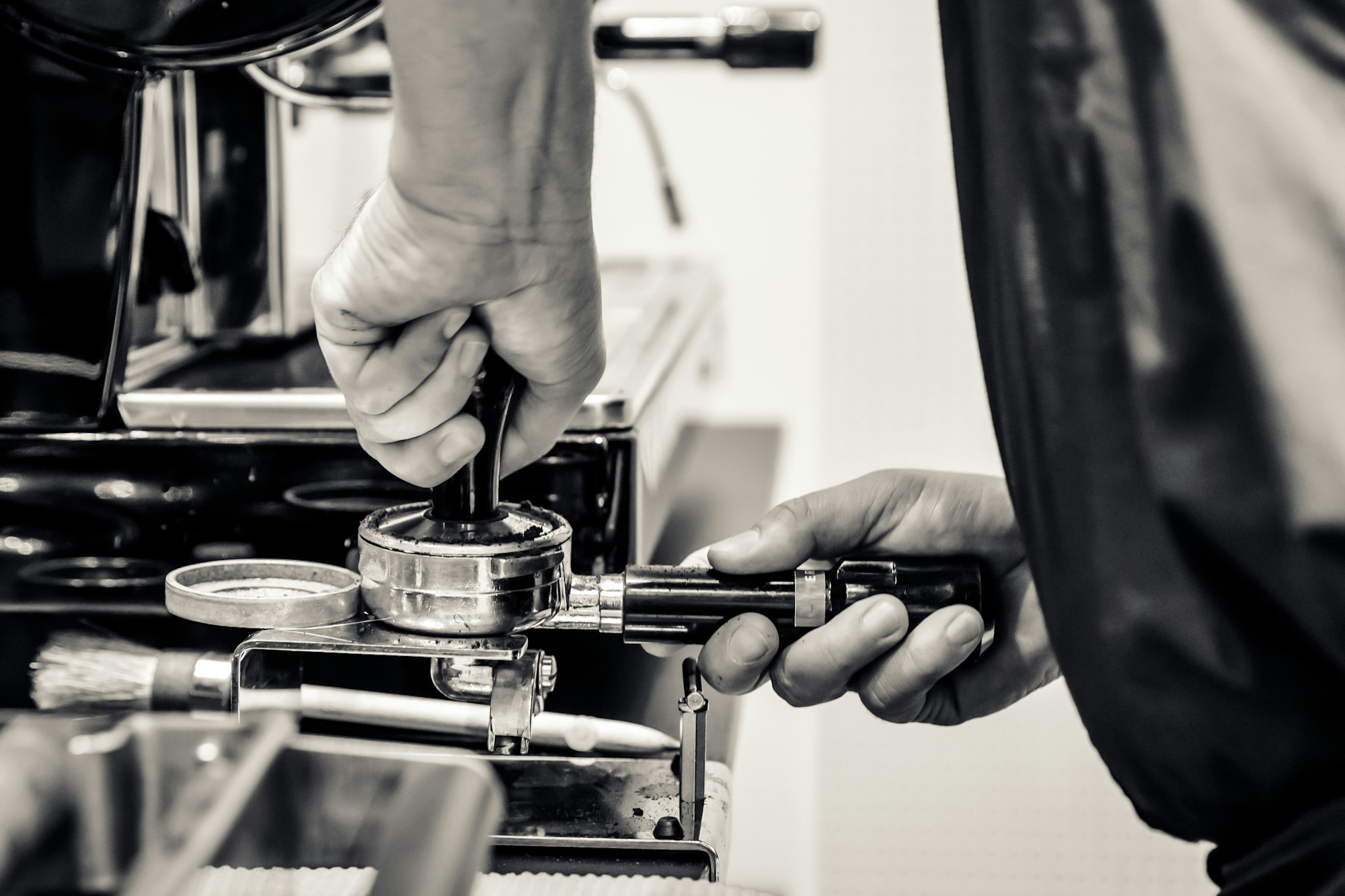 A barista is tamping his coffee at a coffee bar and there is a blurred espresso machine in the background.