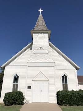 A white wooden church with a simple, traditional design features a prominent steeple topped with a glowing cross. The building's facade includes a pair of tall, narrow windows with pointed arches and a central double door. Bushes are neatly arranged on either side of the entrance, and the sky above is clear and blue.
