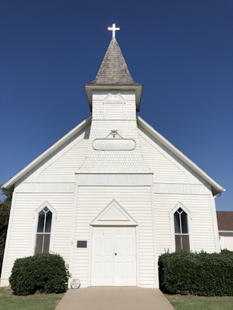 A white wooden church with a simple, traditional design features a prominent steeple topped with a glowing cross. The building's facade includes a pair of tall, narrow windows with pointed arches and a central double door. Bushes are neatly arranged on either side of the entrance, and the sky above is clear and blue.