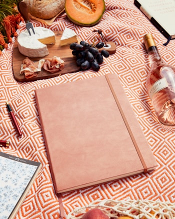 A picnic setup featuring a patterned orange and white blanket. On the blanket are various items including a wooden board with cheese, prosciutto, and grapes. There’s a large notebook or journal, a pen, and a bottle of rosé wine to the side. A halved cantaloupe and a loaf of bread are also present, contributing to a rustic, laid-back vibe.
