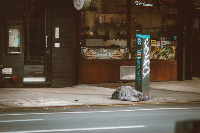 A storefront with a dark exterior, displaying promotional images of food items like quesadillas. The entrance is adjacent to a sidewalk where a pile of clothing or blankets is left on the ground. A parking meter in the foreground is covered in graffiti.