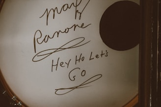 A drum head with handwritten text featuring signatures and phrases, including 'Hey Ho Let's Go'. The surface is predominantly white with a dark circular patch on the right side.