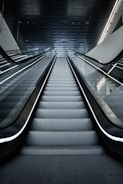 A modern escalator leading upwards, with a sleek and metallic design. The lighting is cool and the setting appears to be indoors, possibly in a mall or a subway station. The ceiling is dark with lighting accents along the sides.