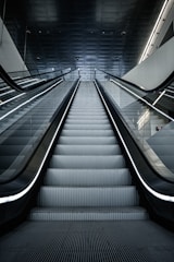 A modern escalator leading upwards, with a sleek and metallic design. The lighting is cool and the setting appears to be indoors, possibly in a mall or a subway station. The ceiling is dark with lighting accents along the sides.