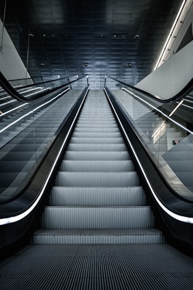 A modern escalator leading upwards, with a sleek and metallic design. The lighting is cool and the setting appears to be indoors, possibly in a mall or a subway station. The ceiling is dark with lighting accents along the sides.