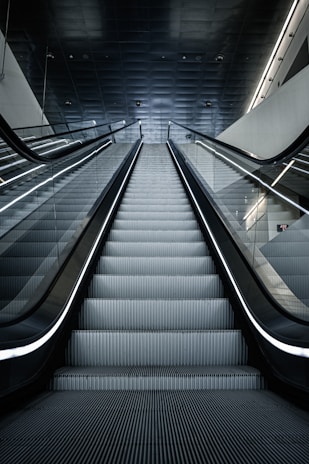 A modern escalator leading upwards, with a sleek and metallic design. The lighting is cool and the setting appears to be indoors, possibly in a mall or a subway station. The ceiling is dark with lighting accents along the sides.