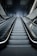 A modern escalator leading upwards, with a sleek and metallic design. The lighting is cool and the setting appears to be indoors, possibly in a mall or a subway station. The ceiling is dark with lighting accents along the sides.