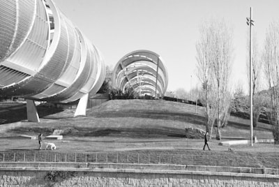 Grayscale photo of round building. People walking dogs under tubular bridge, black and white