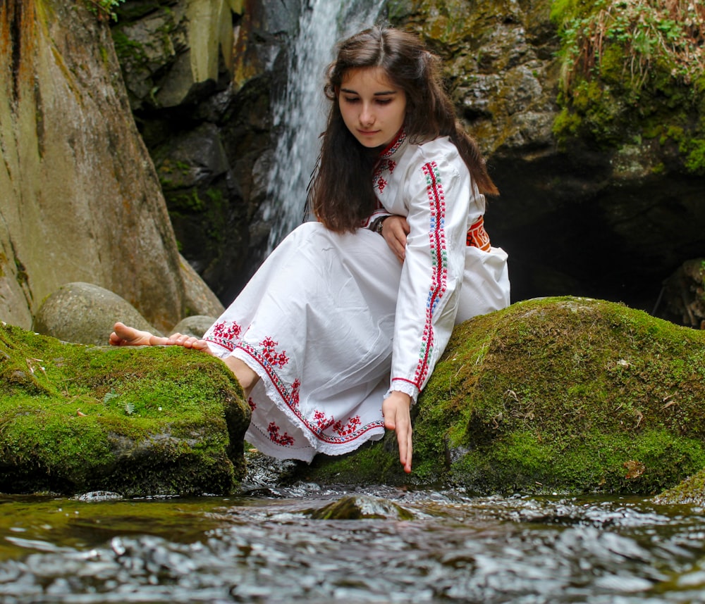 woman in white long sleeve dress sitting on rock by the river during daytime
