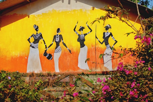 A vibrant mural depicts four stylized figures dressed in traditional clothing and adorned with jewelry. The art uses black and white against a bright orange and yellow gradient background. Nearby, colorful pink and purple flowers add to the scene, along with green foliage in the foreground.