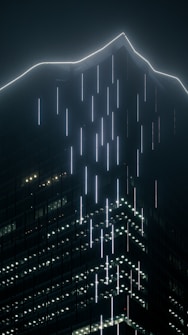 A towering skyscraper with a sleek, modern design is illuminated by vertical lines of bright white lights against a dark, foggy background. The top of the building features angular, sharp lines that give it a futuristic appearance.