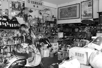 A cluttered and busy garage or workshop filled with various tools, equipment, and boxes. Shelves are stocked with helmets, spray cans, and other motorcycle-related products. A scooter is parked in the foreground, and a fan is mounted on the wall. Posters and signs decorate the walls, adding to the chaotic yet functional atmosphere.