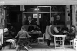 A group of people sit outside a small, casual restaurant or cafe. Several individuals are seated at tables, engaged in conversation or quietly enjoying their surroundings. The area appears somewhat cluttered with tables and chairs, and the location seems to be in an urban setting. The atmosphere suggests a communal and relaxed environment with a local charm, indicated by the informal arrangement and variety of patrons.