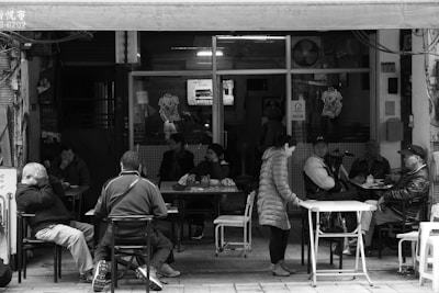 A group of people sit outside a small, casual restaurant or cafe. Several individuals are seated at tables, engaged in conversation or quietly enjoying their surroundings. The area appears somewhat cluttered with tables and chairs, and the location seems to be in an urban setting. The atmosphere suggests a communal and relaxed environment with a local charm, indicated by the informal arrangement and variety of patrons.