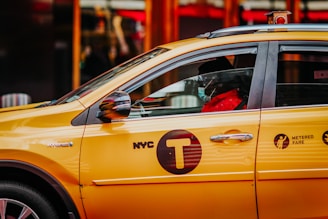 A yellow New York City taxi is parked with a visible driver wearing a mask inside. The taxi has visible 'NYC T' and 'Metered Fare' signage on its side. The image captures the side view of the car.