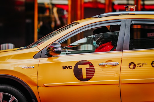 A yellow New York City taxi is parked with a visible driver wearing a mask inside. The taxi has visible 'NYC T' and 'Metered Fare' signage on its side. The image captures the side view of the car.