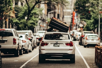 A white BMW SUV is parked in the middle of a city street with a large couch strapped to its roof. The surrounding area is filled with other vehicles lined up along the road, indicative of an urban setting. The foliage of trees lines the street, suggesting a blend of city and nature.