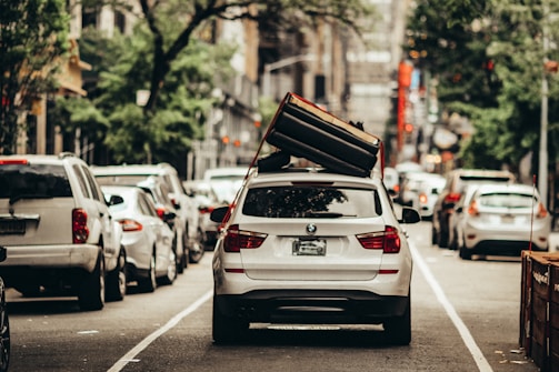 A white BMW SUV is parked in the middle of a city street with a large couch strapped to its roof. The surrounding area is filled with other vehicles lined up along the road, indicative of an urban setting. The foliage of trees lines the street, suggesting a blend of city and nature.
