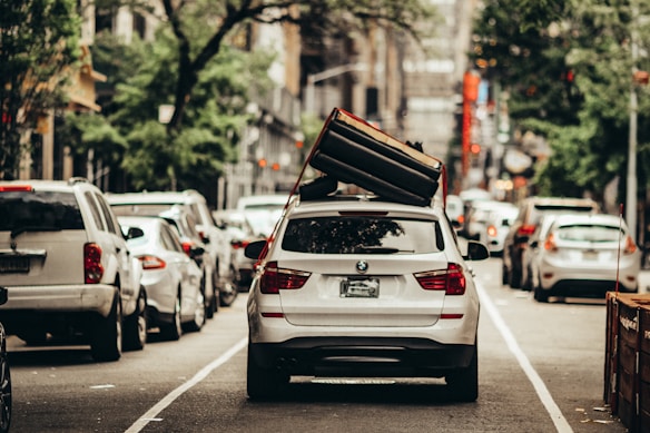 A white BMW SUV is parked in the middle of a city street with a large couch strapped to its roof. The surrounding area is filled with other vehicles lined up along the road, indicative of an urban setting. The foliage of trees lines the street, suggesting a blend of city and nature.