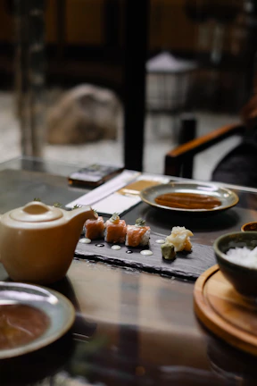 A table set with a slate serving board containing sushi rolls, wasabi, and pickled ginger. There is a ceramic teapot and plates on the glass table. The setting appears elegant and modern, with soft lighting creating a cozy atmosphere.