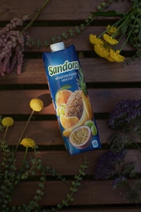 A carton of multivitamin nectar with images of various fruits like pineapple, orange, and passion fruit on it. It is placed on a wooden surface surrounded by an arrangement of colorful flowers, including yellow, purple, and pink variations.