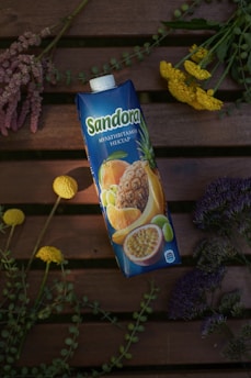 A carton of multivitamin nectar with images of various fruits like pineapple, orange, and passion fruit on it. It is placed on a wooden surface surrounded by an arrangement of colorful flowers, including yellow, purple, and pink variations.