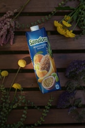 A carton of multivitamin nectar with images of various fruits like pineapple, orange, and passion fruit on it. It is placed on a wooden surface surrounded by an arrangement of colorful flowers, including yellow, purple, and pink variations.