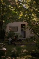 A cozy caravan surrounded by lush green foliage with string lights hanging above. A wooden deck is visible in front of the caravan, adorned with a chair draped with a green blanket. A large lantern and some plants enhance the natural setting.