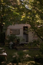A cozy caravan surrounded by lush green foliage with string lights hanging above. A wooden deck is visible in front of the caravan, adorned with a chair draped with a green blanket. A large lantern and some plants enhance the natural setting.