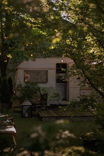 A cozy caravan surrounded by lush green foliage with string lights hanging above. A wooden deck is visible in front of the caravan, adorned with a chair draped with a green blanket. A large lantern and some plants enhance the natural setting.
