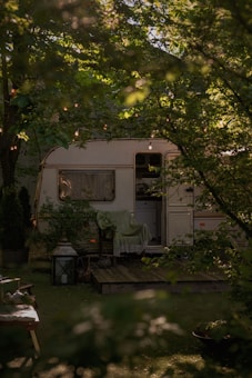 A cozy caravan surrounded by lush green foliage with string lights hanging above. A wooden deck is visible in front of the caravan, adorned with a chair draped with a green blanket. A large lantern and some plants enhance the natural setting.