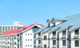 A series of modern buildings with sloped roofs in light grey and red tones under a clear blue sky. The structures have multiple rows of windows and appear to be residential or commercial properties.