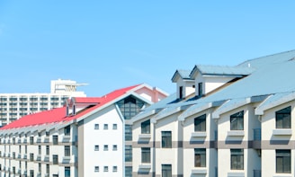 A series of modern buildings with sloped roofs in light grey and red tones under a clear blue sky. The structures have multiple rows of windows and appear to be residential or commercial properties.