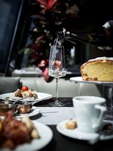 A dining table is set with plates of gourmet food, including dishes with vegetables and a slice of cake on a stand. A gloved hand is pouring a drink from a bottle into an elegant glass. In the background, there are vibrant red and green leaves from a plant.