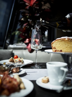 A dining table is set with plates of gourmet food, including dishes with vegetables and a slice of cake on a stand. A gloved hand is pouring a drink from a bottle into an elegant glass. In the background, there are vibrant red and green leaves from a plant.
